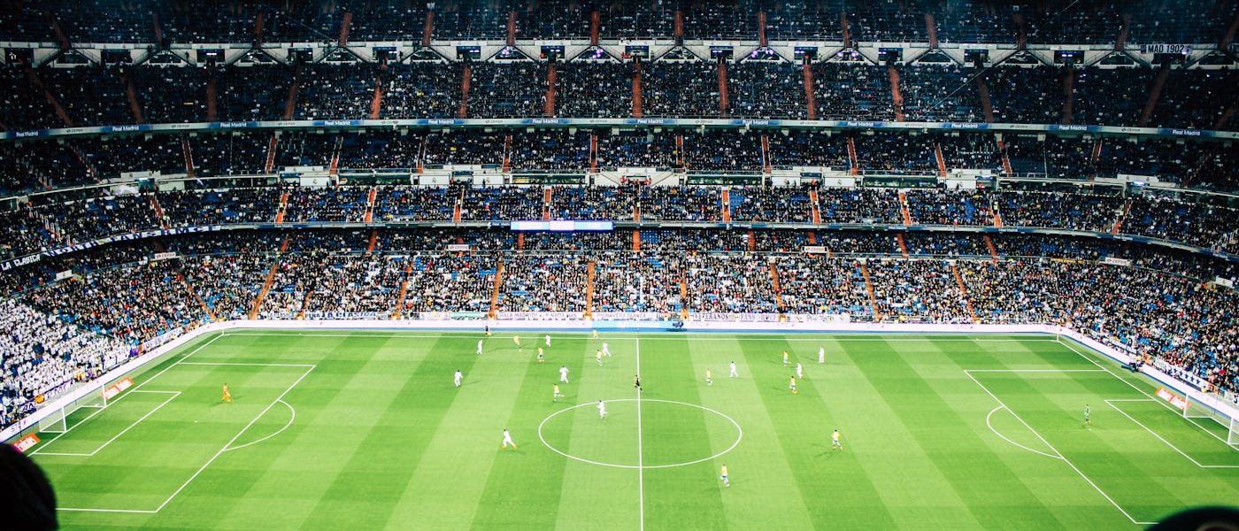 Aerial view of a packed football stadium at night with players on a bright green pitch and illuminated stands above the field.